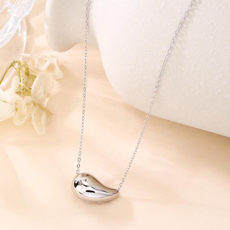 May include: A silver-colored necklace with a teardrop-shaped pendant. The pendant is smooth and reflective, hanging from a delicate chain. The necklace is displayed against a soft, neutral background, highlighting its minimalist design.