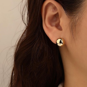 May include: A close-up of a gold-coloured, spherical stud earring. The earring is smooth and reflective, catching the light. The earring is worn on a person's ear, with dark brown hair visible in the background. The earring is a simple, classic design.