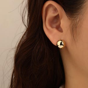 May include: A close-up of a gold-coloured, spherical stud earring. The earring is a smooth, polished sphere, reflecting light. The earring is worn on an ear, with the person's hair and skin visible in the background. The earring is a simple, elegant design.