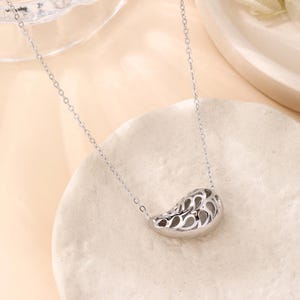 May include: A silver-colored necklace with a bean-shaped pendant. The pendant has a hollow, openwork design with a repeating pattern. The necklace chain is delicate and thin. The jewelry is displayed on a light-colored surface.