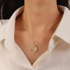 May include: A gold-colored necklace featuring a crescent moon pendant suspended from a circular and straight bar design. The necklace is worn against a white shirt, highlighting the jewelry's minimalist style and elegant aesthetic.