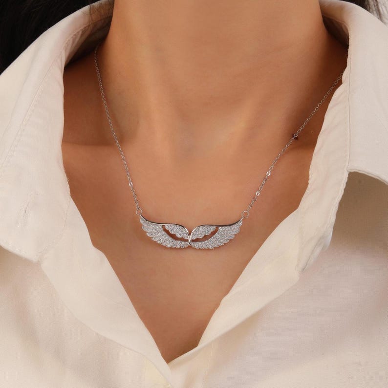 May include: A silver necklace featuring a pair of stylized angel wings, encrusted with small, sparkling stones. The wings are positioned horizontally, with the tips curving slightly upwards. The necklace is worn against a cream-colored shirt.