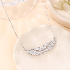 Op de afbeelding: Een zilverkleurige ketting met een paar gestileerde vleugels, bezet met kleine, sprankelende steentjes. De vleugels zijn het centrale punt, met een fijne ketting die zich aan weerszijden uitstrekt. De ketting ligt op een lichtgekleurd, getextureerd oppervlak.