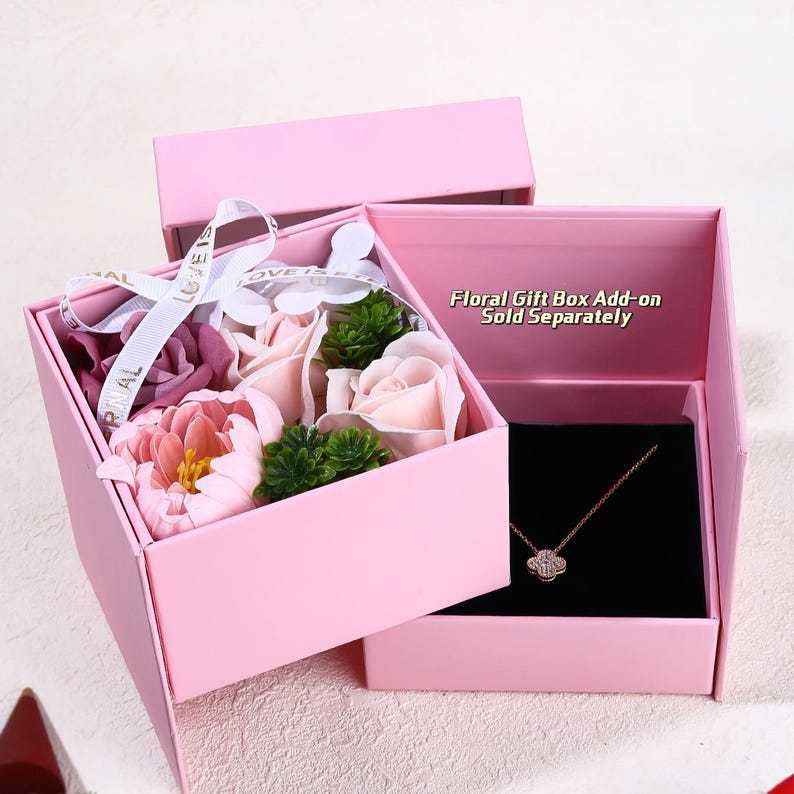 May include: A pink gift box containing a floral arrangement of pink and purple roses, and green succulents. A white ribbon with the text "LOVE IS ETERNAL" is tied around the flowers. A gold necklace with a clover pendant rests on a black velvet insert.