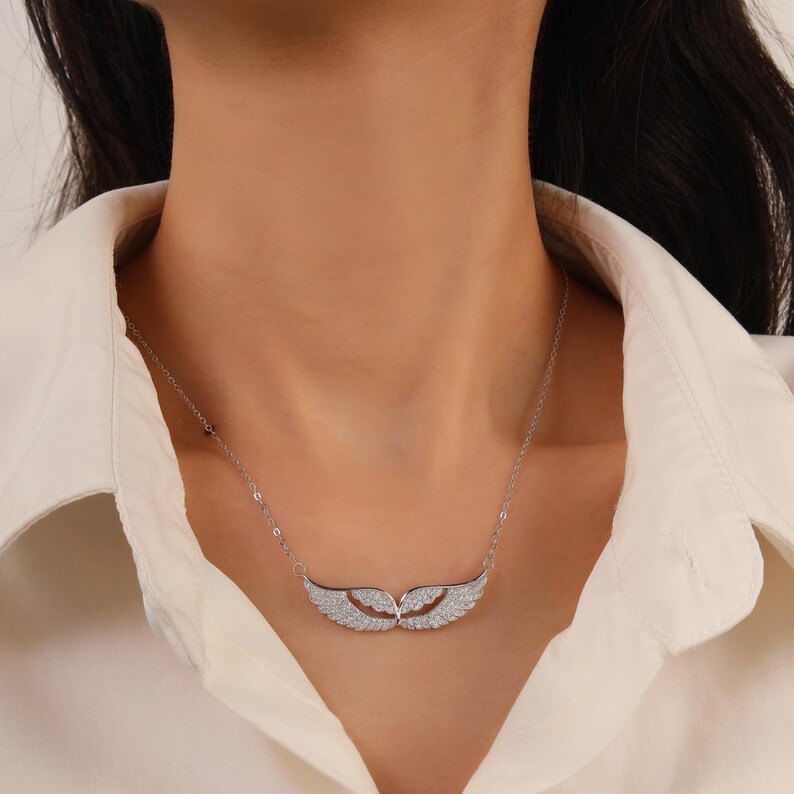 May include: A silver necklace featuring a pair of stylized wings, encrusted with small, sparkling stones. The wings are positioned horizontally, connected by a delicate chain. The necklace is worn against a white shirt.