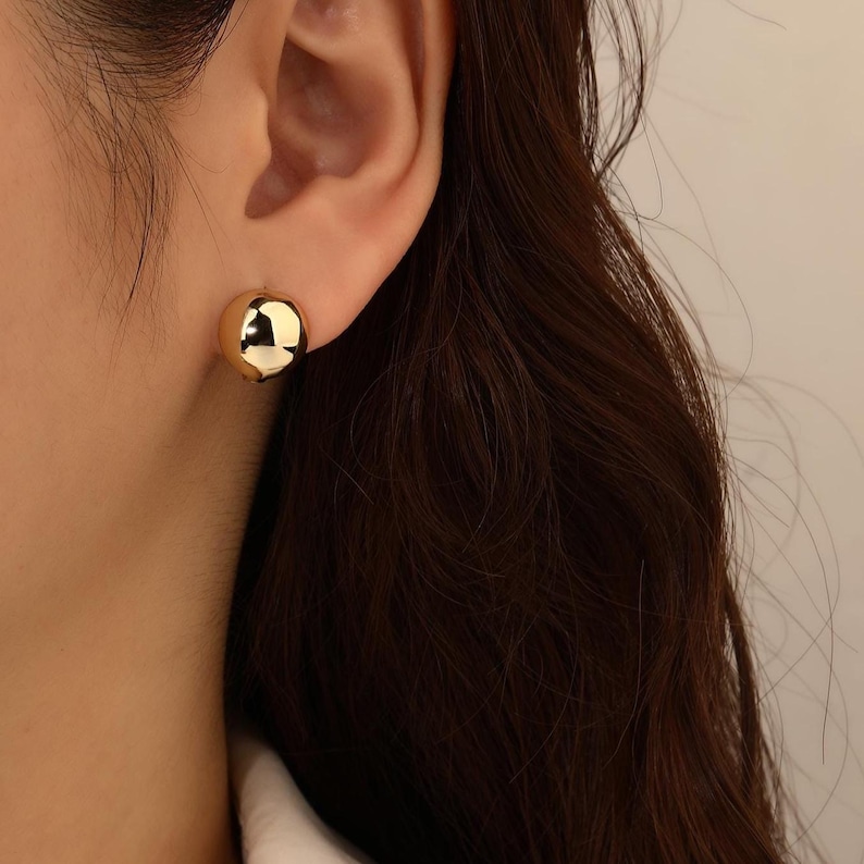 May include: A close-up of a gold-coloured, spherical stud earring. The earring is a smooth, polished sphere, reflecting light. The earring is worn on an ear, with dark hair visible in the background. The earring is a simple, elegant accessory.