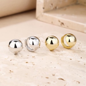 May include: Two pairs of ball-shaped hoop earrings, one silver and one gold. The earrings are presented on a light, textured surface. The earrings have a polished, reflective finish.