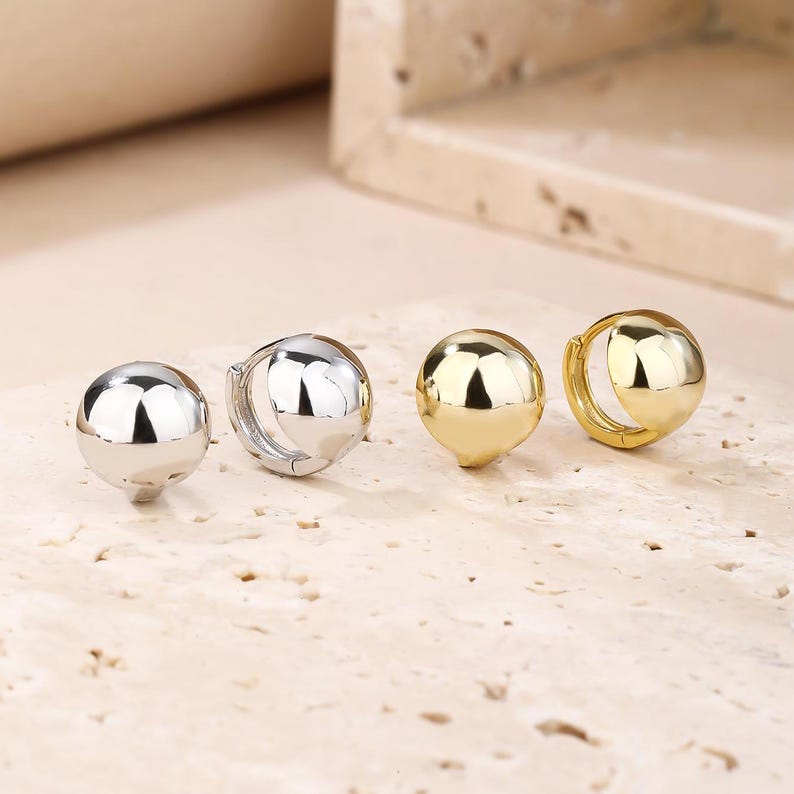 May include: Two pairs of ball-shaped hoop earrings, one silver and one gold. The earrings are presented on a light, textured surface. The earrings have a polished, reflective finish.