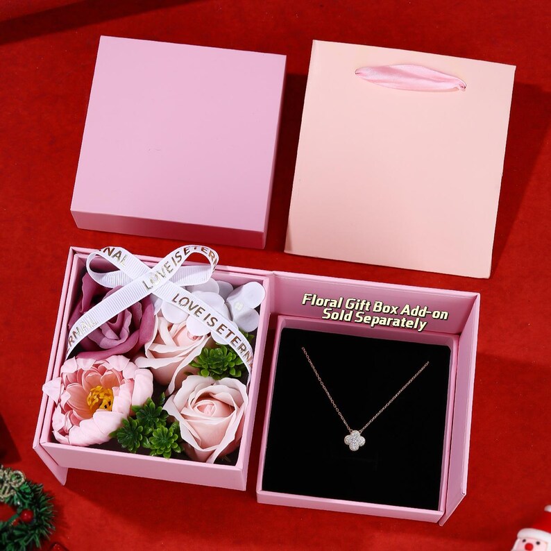 May include: A pink gift box containing a floral arrangement and a necklace with a four-leaf clover pendant. The box has a ribbon that reads "LOVE IS ETERNAL". A matching pink gift bag is also present. The text "Floral Gift Box Add-on Sold Separately" is visible.