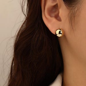 May include: A close-up of a gold-coloured, spherical stud earring. The earring is smooth and reflective, catching the light. The earring is worn on the ear of a person with dark brown hair.