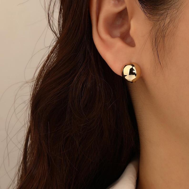 May include: A close-up of a gold-coloured, spherical stud earring. The earring is smooth and reflective, catching the light. The earring is worn on the ear of a person with dark brown hair.