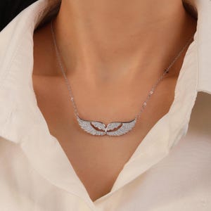May include: A silver necklace featuring a pair of stylized wings, encrusted with small, sparkling stones. The wings are connected by a delicate chain, creating a graceful and elegant design. The necklace is worn against a white shirt.