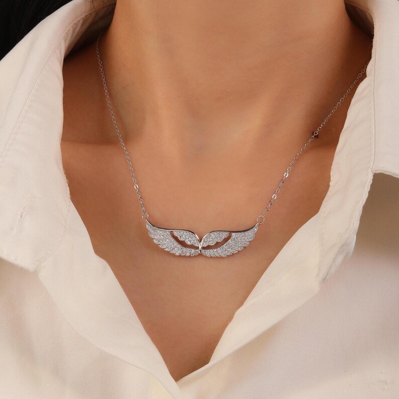 May include: A silver necklace featuring a pair of stylized wings, encrusted with small, sparkling stones. The wings are connected by a delicate chain, creating a graceful and elegant design. The necklace is worn against a white shirt.