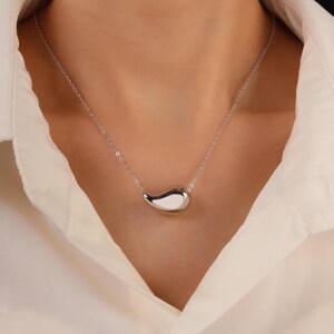 May include: A silver-colored necklace featuring a teardrop-shaped pendant. The pendant is smooth and reflective, hanging from a delicate chain. The necklace is displayed against a white shirt.