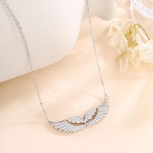 May include: A silver-colored necklace featuring a wing design. The pendant is adorned with small, sparkling stones, creating a shimmering effect. The necklace hangs from a delicate chain, showcasing a graceful and elegant style.