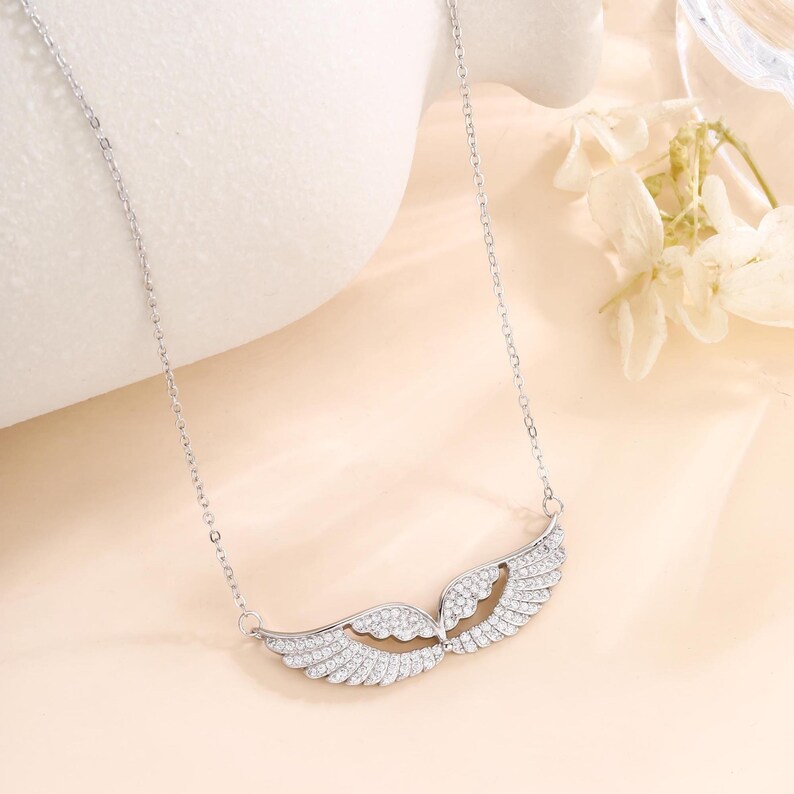 May include: A silver-colored necklace featuring a wing design. The pendant is adorned with small, sparkling stones, creating a shimmering effect. The necklace hangs from a delicate chain, showcasing a graceful and elegant style.