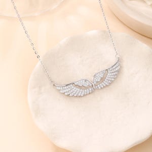May include: A silver necklace featuring a pair of angel wings, encrusted with small, sparkling stones. The wings are detailed with individual feather designs and are connected by a delicate chain. The necklace is displayed on a light-colored, textured surface.
