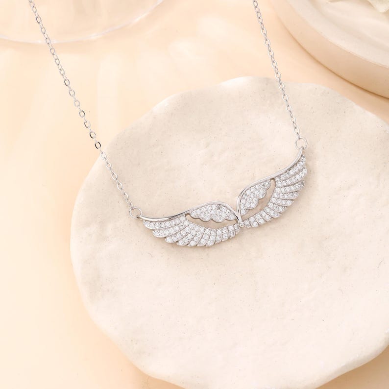 May include: A silver necklace featuring a pair of angel wings, encrusted with small, sparkling stones. The wings are detailed with individual feather designs and are connected by a delicate chain. The necklace is displayed on a light-colored, textured surface.