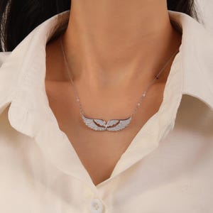 May include: A silver necklace featuring a pair of stylized wings, encrusted with small, sparkling stones. The wings are positioned horizontally, suspended from a delicate chain. The necklace is worn against a white collared shirt.