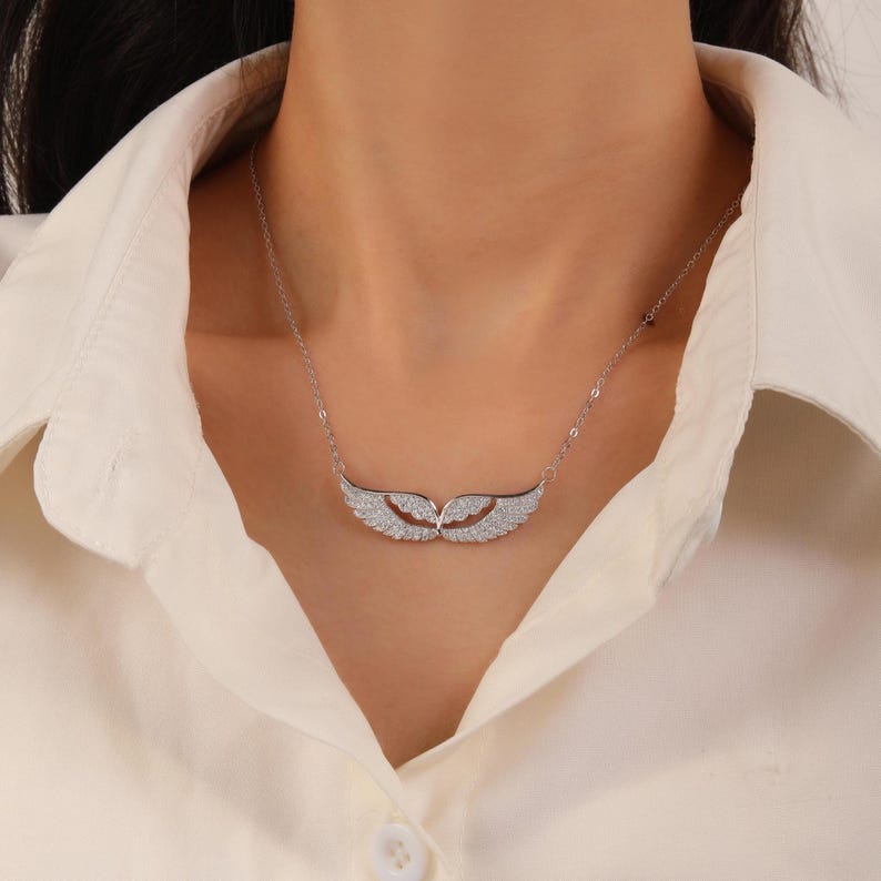May include: A silver necklace featuring a pair of stylized wings, encrusted with small, sparkling stones. The wings are positioned horizontally, suspended from a delicate chain. The necklace is worn against a white collared shirt.