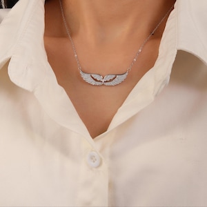 May include: A silver necklace featuring a pendant shaped like a pair of wings, encrusted with small, sparkling stones. The necklace is worn against a cream-colored collared shirt, creating a contrast. The wings are approximately 1.5 inches wide.