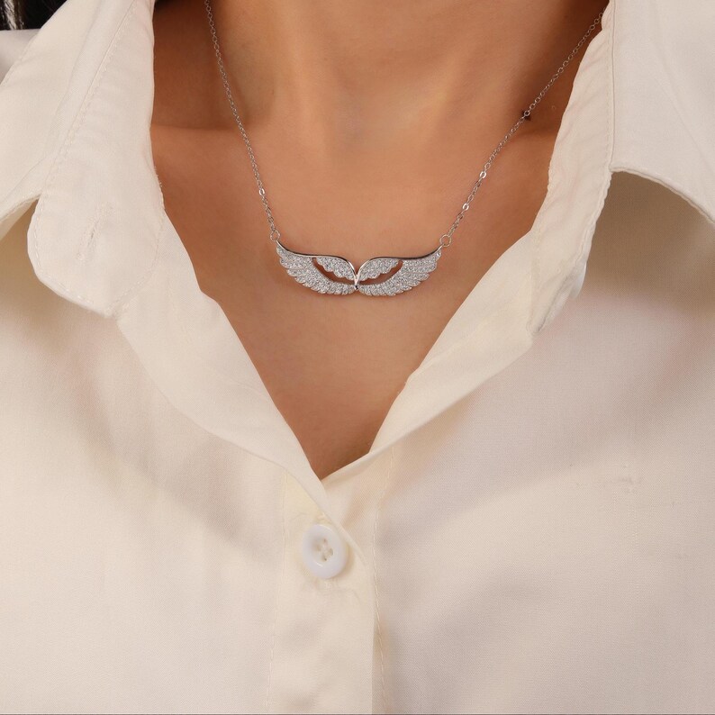 May include: A silver necklace featuring a pendant shaped like a pair of wings, encrusted with small, sparkling stones. The necklace is worn against a cream-colored collared shirt, creating a contrast. The wings are approximately 1.5 inches wide.