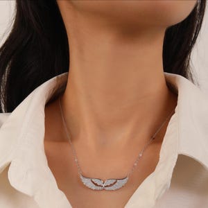 May include: A silver necklace featuring a pendant shaped like angel wings. The wings are encrusted with small, sparkling stones. The necklace is worn against a white shirt.
