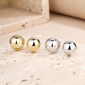 May include: Four pairs of half-sphere earrings. Two pairs are gold-toned, and two are silver-toned. The earrings have a polished, reflective surface. The earrings are displayed on a light-coloured, textured surface.