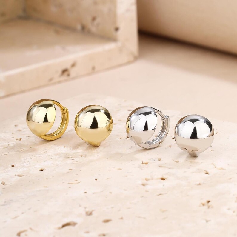 May include: Four pairs of half-sphere earrings. Two pairs are gold-toned, and two are silver-toned. The earrings have a polished, reflective surface. The earrings are displayed on a light-coloured, textured surface.