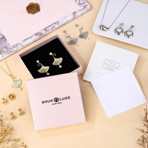 May include: A collection of jewelry, including gold and silver earrings, necklaces, and studs. The earrings and necklace feature a leaf design with a pearl accent. The jewelry is displayed in a pink box with the brand name "SHUK LUXE NEW YORK".