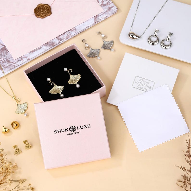 May include: A collection of jewelry, including gold and silver earrings, necklaces, and studs. The earrings and necklace feature a leaf design with a pearl accent. The jewelry is displayed in a pink box with the brand name "SHUK LUXE NEW YORK".