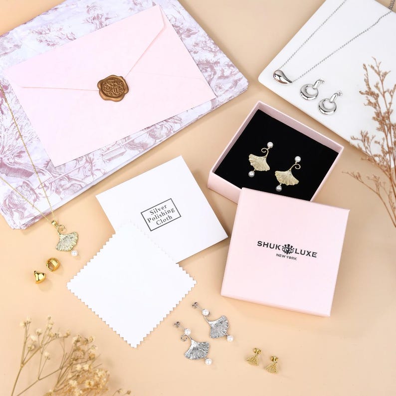 May include: An overhead shot of jewellery and accessories. A pink box with the text "SHUK LUXE NEW YORK" contains gold leaf-shaped earrings with pearls. Other jewellery includes silver necklaces and earrings, and gold stud earrings. A pink envelope is sealed with a gold wax stamp.