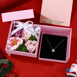 May include: A pink gift box with a floral arrangement and a necklace. The box contains a necklace with a four-leaf clover pendant and a ribbon that says "LOVE IS ETERNAL". The text "Floral Gift Box Add-on Sold Separately" is also visible.