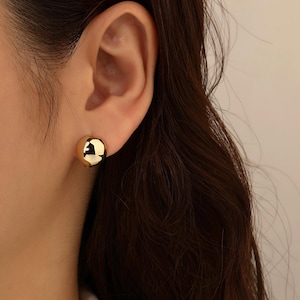 May include: A close-up of a gold-coloured, spherical stud earring. The earring is smooth and reflective, catching the light. The earring is worn on an ear, with dark brown hair visible.