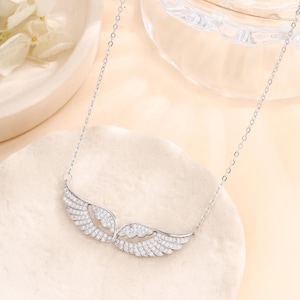 May include: A silver necklace featuring a pair of open wings, encrusted with small, sparkling stones. The wings are the focal point, suspended from a delicate chain. The necklace is displayed on a neutral-toned surface.