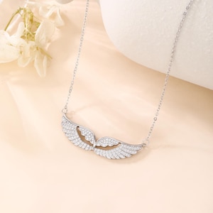 May include: A silver-colored necklace featuring a pair of open wings, encrusted with small, sparkling crystals. The wings are the focal point, suspended from a delicate chain. The background is a soft, neutral color, highlighting the jewelry.