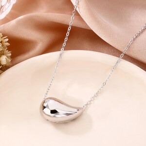 May include: A silver-colored necklace with a bean-shaped pendant. The pendant is smooth and reflective, catching the light. The chain is delicate and links to the pendant. The necklace is displayed on a white plate and a peach-colored fabric.