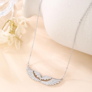 May include: A silver necklace featuring a pair of angel wings, encrusted with sparkling clear crystals. The wings are detailed with individual feather designs and are suspended from a delicate chain. The necklace is displayed on a light peach background.