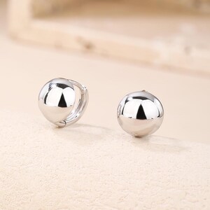 May include: A pair of shiny, silver-coloured ball hoop earrings. The earrings have a smooth, reflective surface, and the round shape is a classic design. The earrings are shown on a neutral background.