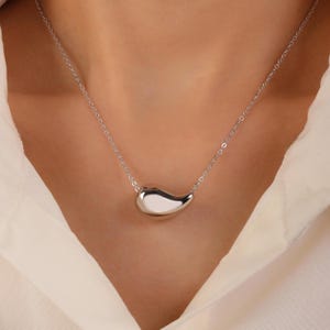 May include: A silver necklace featuring a teardrop-shaped pendant. The pendant is smooth and reflective, suspended from a delicate chain. The necklace is worn against a white shirt.