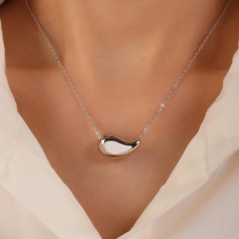 May include: A silver necklace featuring a teardrop-shaped pendant. The pendant is smooth and reflective, suspended from a delicate chain. The necklace is worn against a white shirt.