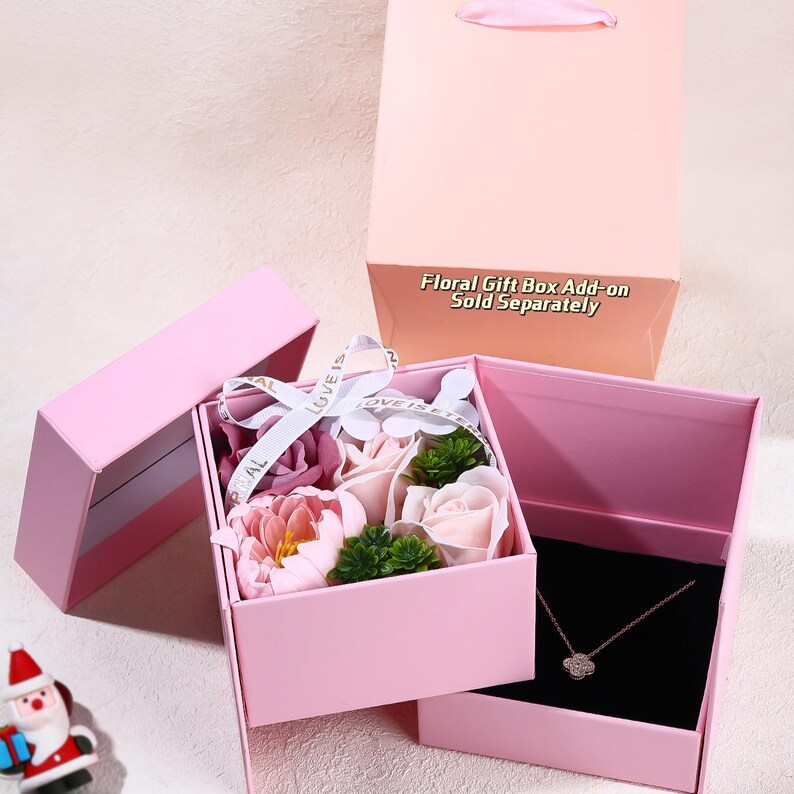 May include: A pink gift box with a white ribbon that reads "Love is". The box contains artificial flowers and a necklace. A pink gift bag is in the background. The text "Floral Gift Box Add-on Sold Separately" is visible.