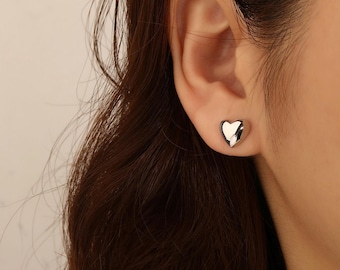 May include: A small, polished silver-tone heart-shaped stud earring with a smooth, reflective surface. The earring is secured to the earlobe, featuring a rounded, three-dimensional design that catches the light, creating bright highlights.