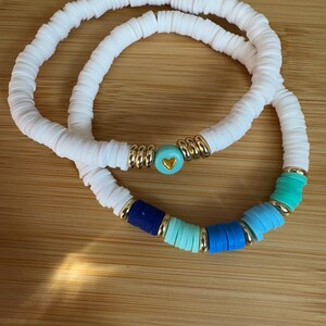 Op de afbeelding: Twee witte kralen armbanden met een gouden hartbedel en blauwe en turquoise kralen.