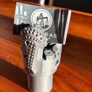 May include: A grey 3D-printed business card holder shaped like a tri-cone drill bit. The holder features a textured, metallic-looking finish and holds a rectangular card with a circular logo depicting an oil derrick and the text GAS.