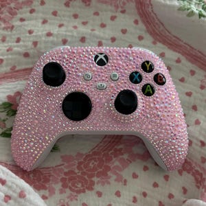 May include: An Xbox wireless controller fully encrusted with small, light pink iridescent rhinestones. The controller features two black analog sticks, a black directional pad, and standard white buttons with the Xbox logo and colored A, B, X, Y labels.
