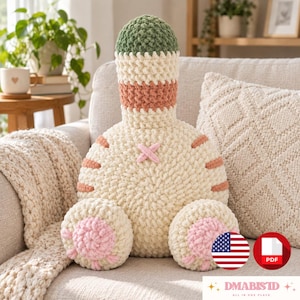May include: A crochet plush pillow shaped like a cat rear, featuring a cream-colored body, a green and cream striped tail, pink paw pads on two round feet, and a small pink cross detail. The texture is thick, soft, and yarn-based throughout.