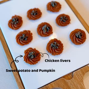 May include: A white ceramic serving tray with a wooden frame, holding eight small, round, orange-brown sweet potato and pumpkin treats with a dark brown chicken liver topping. The text "Sweet potato and Pumpkin" and "Chicken livers" is written on the tray.