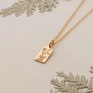 May include: A delicate gold-colored necklace with a rectangular pendant featuring a floral design. The necklace rests on a white surface, adorned with dried fern leaves. The pendant is approximately 0.5 inches in length.