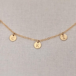 May include: A gold chain necklace with five small gold discs hanging from it. Each disc has a letter stamped on it, spelling out the word 'LAKE'. The necklace is on a white fabric background.